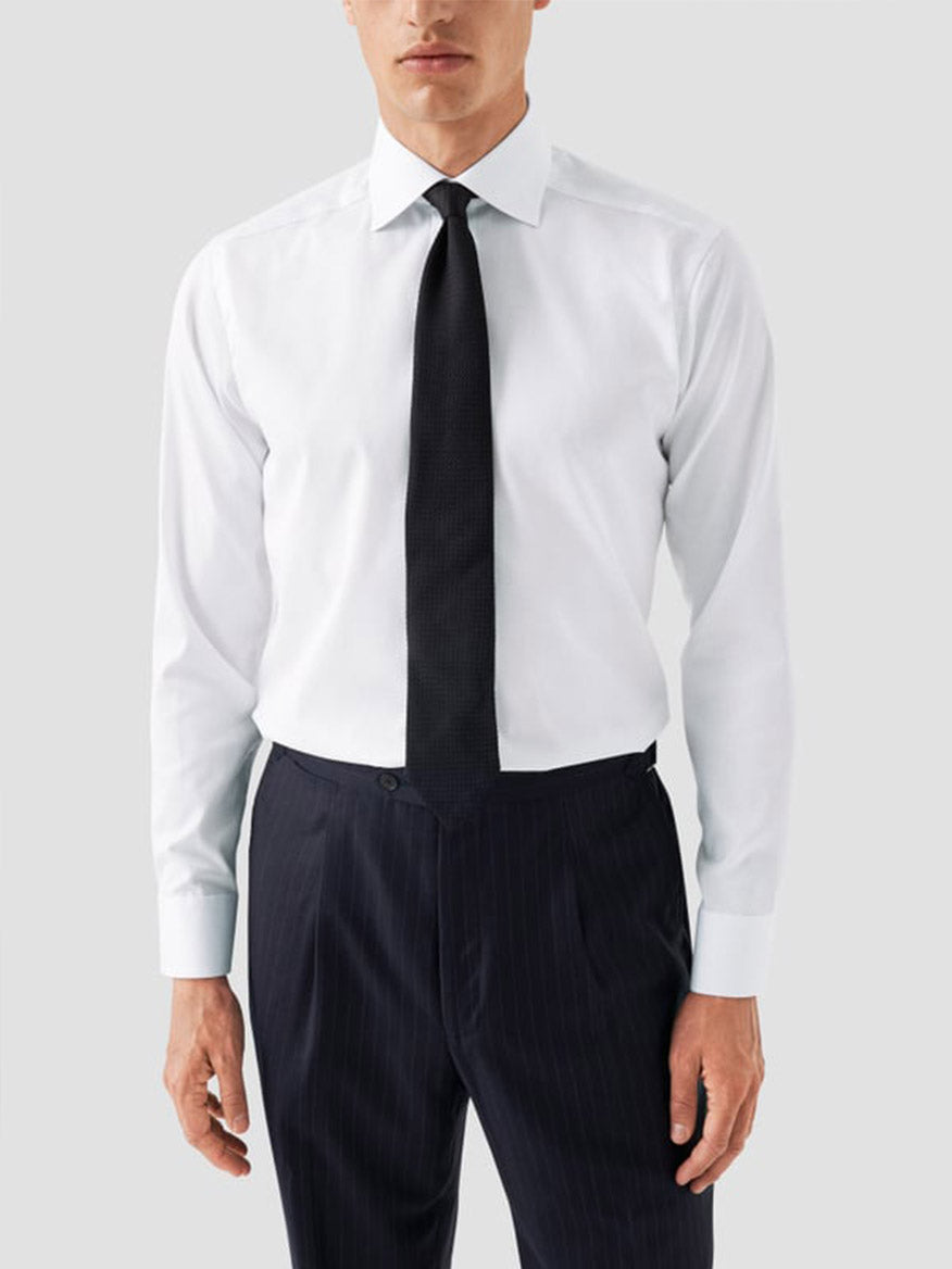 A man wearing the Eton Slim Fit Solid Signature Twill Dress Shirt in white with navy details, paired with a black tie and dark pinstripe trousers, stands against a plain light gray background.