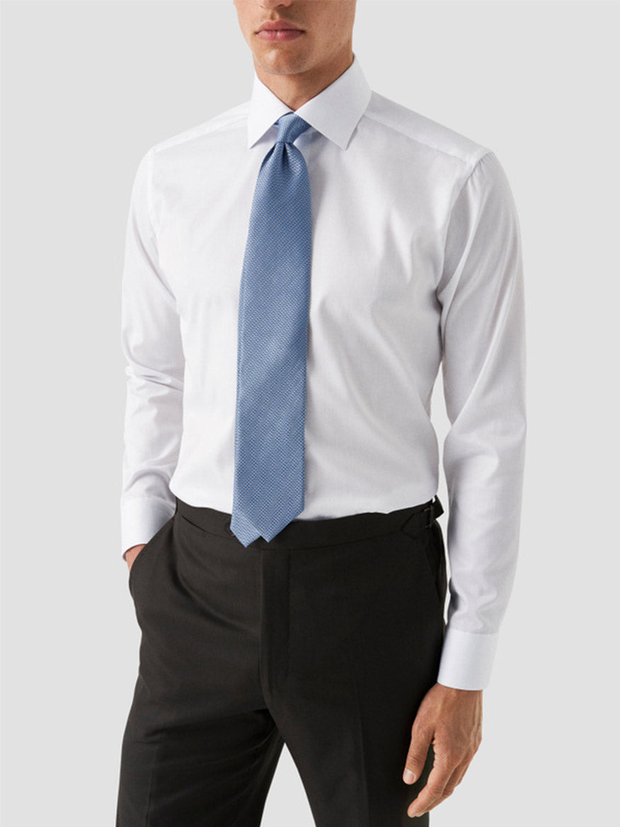 A man wears the Eton Contemporary Fit White Twill Dress Shirt with blue details, paired with a blue tie and black dress pants, standing with one hand in his pocket against a plain light background for a modern look.