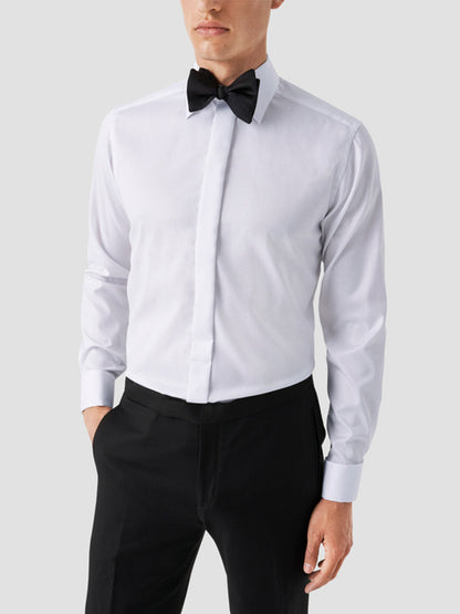 A man wears the Eton Slim Fit Fly Front Signature Twill Evening Shirt in white with a black bow tie and black pants, standing against a plain light gray background.