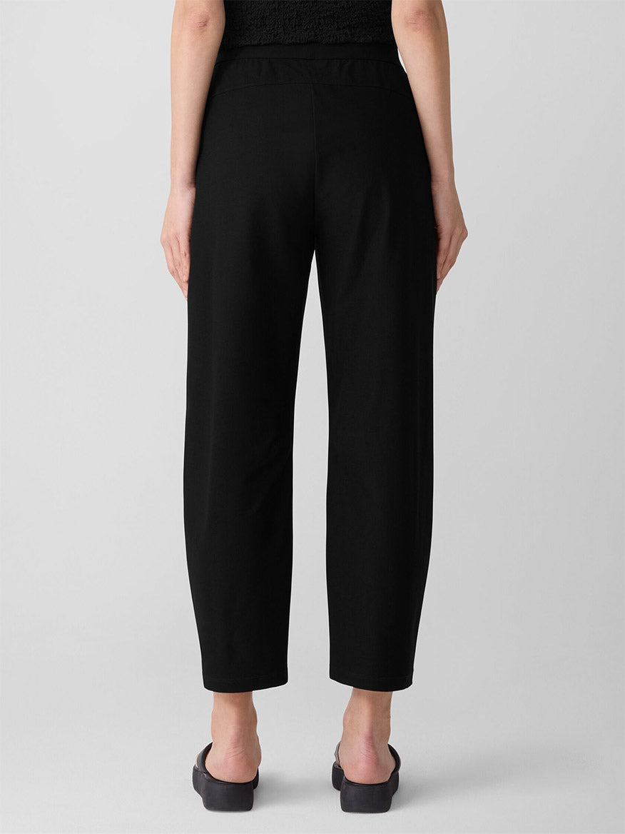 A person faces away from the camera, wearing the Eileen Fisher Washable Stretch Crepe Arch Pant in Black and black slip-on shoes, set against a plain background.