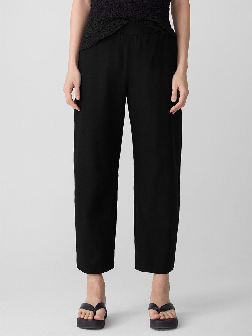 A person stands against a plain light background wearing the Eileen Fisher Washable Stretch Crepe Arch Pant in Black, styled with a black top and black flip-flops.