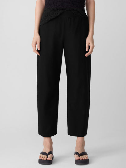 A person stands against a plain light background wearing the Eileen Fisher Washable Stretch Crepe Arch Pant in Black, styled with a black top and black flip-flops.