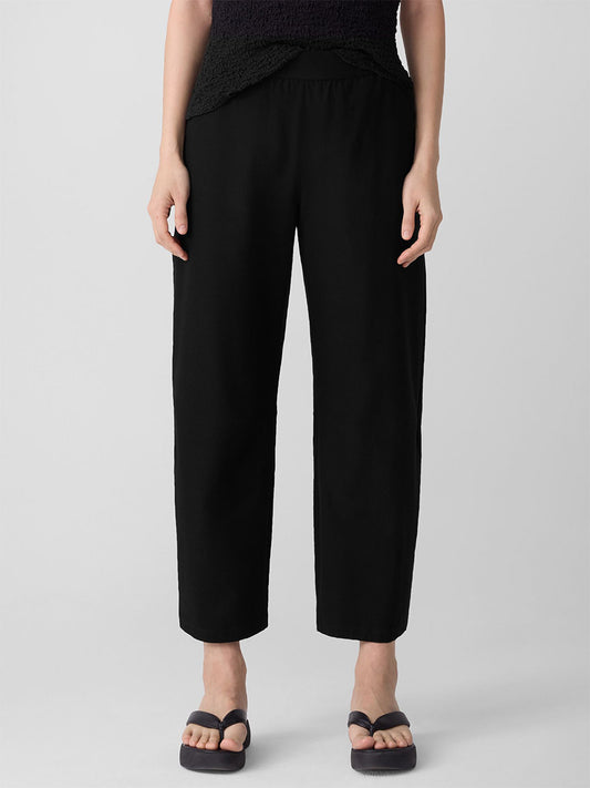 A person stands against a plain light background wearing the Eileen Fisher Washable Stretch Crepe Arch Pant in Black, styled with a black top and black flip-flops.