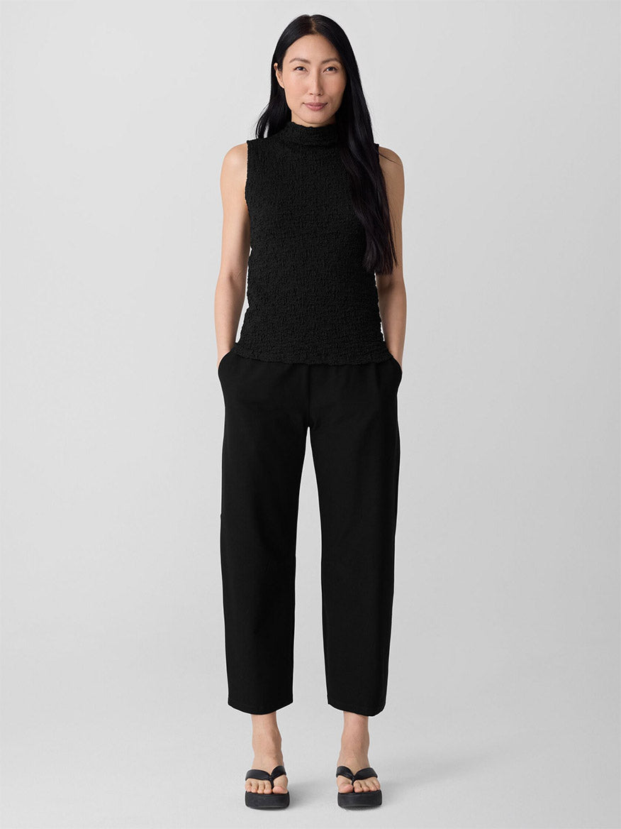 A woman stands against a plain background wearing the Eileen Fisher Washable Stretch Crepe Arch Pant in Black, paired with a sleeveless black top and black sandals, her hands resting in her pockets.