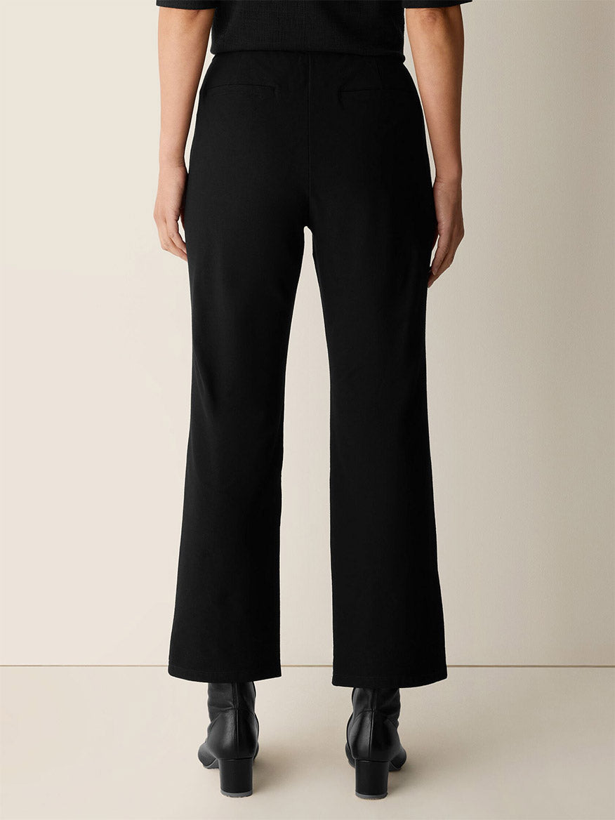 Person standing with their back to the camera, wearing Eileen Fisher Washable Stretch Crepe Kick Pant in Black, styled with ankle boots against a plain background.