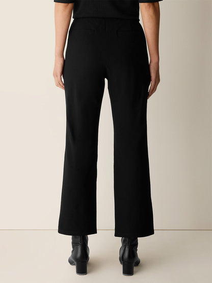 Person standing with their back to the camera, wearing Eileen Fisher Washable Stretch Crepe Kick Pant in Black, styled with ankle boots against a plain background.