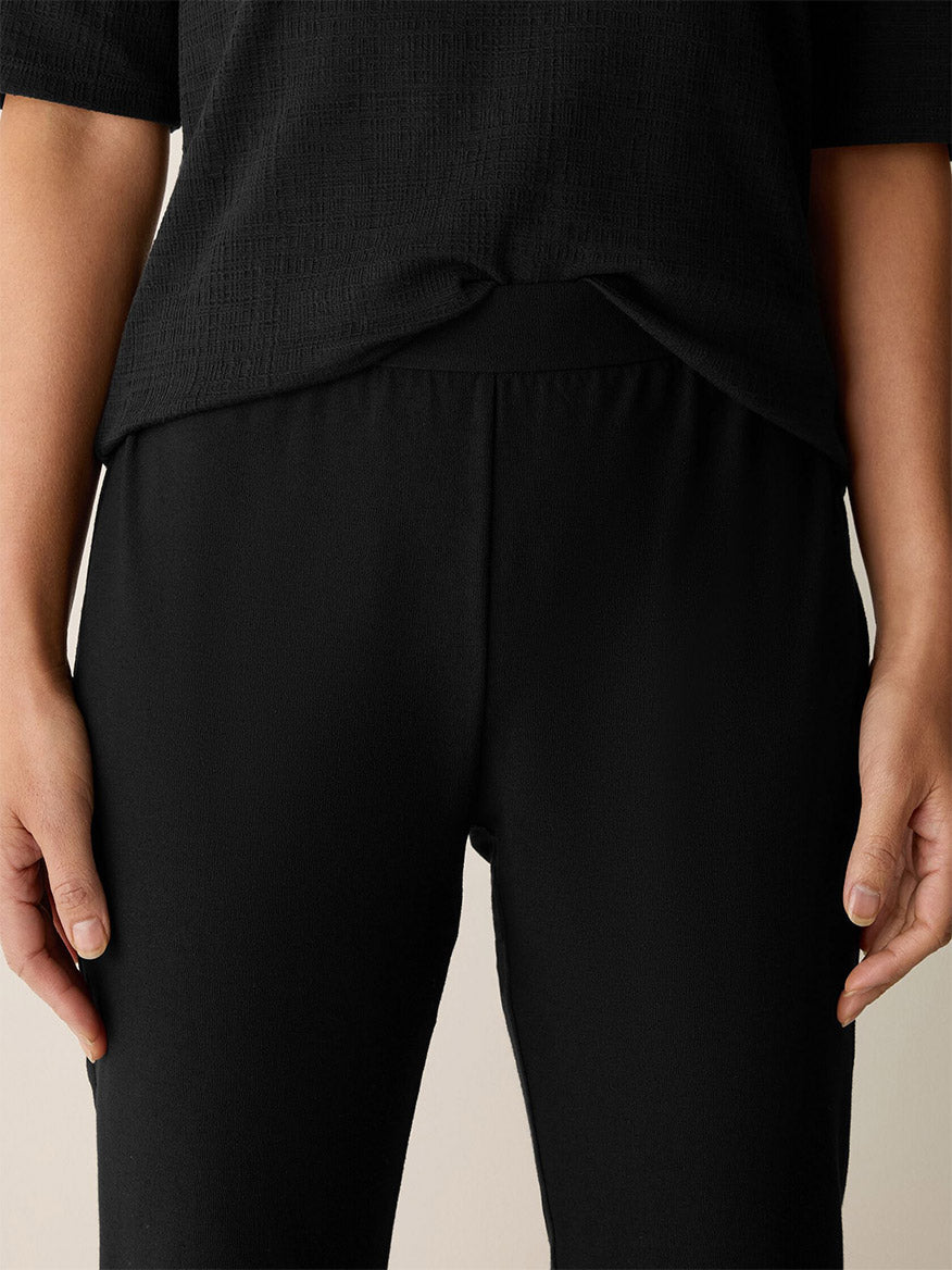 Someone wears the Eileen Fisher Washable Stretch Crepe Kick Pant in Black, shown from waist to mid-thigh, with hands at their sides.