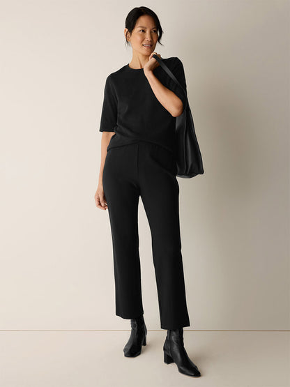 A woman stands against a plain background, dressed in a black short-sleeved top, Eileen Fisher Washable Stretch Crepe Kick Pant in Black, black ankle boots, and carrying a black shoulder bag.