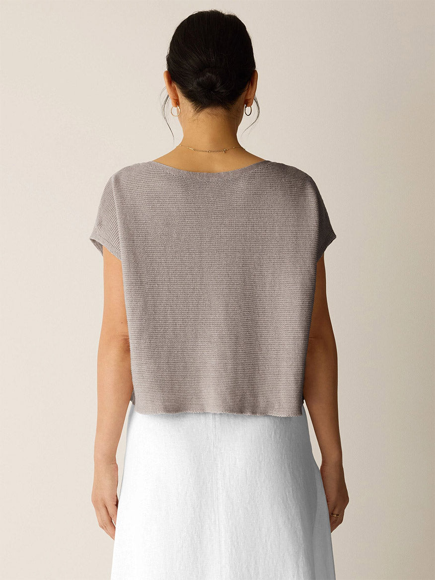 A woman faces away, wearing the Eileen Fisher Organic Linen Délavé Square Top in Walnut—a breathable, boxy-fit taupe knit with short sleeves—paired with a white skirt against a neutral backdrop.