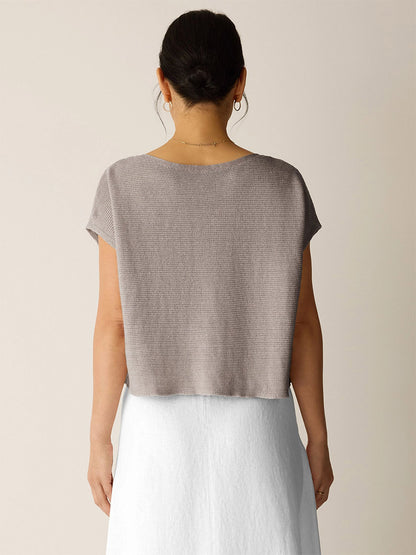 A woman faces away, wearing the Eileen Fisher Organic Linen Délavé Square Top in Walnut—a breathable, boxy-fit taupe knit with short sleeves—paired with a white skirt against a neutral backdrop.