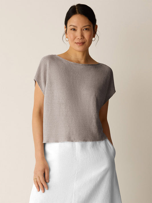 A woman faces forward in the Eileen Fisher Organic Linen Délavé Square Top in Walnut—a breathable, boxy-fit taupe knit—paired with a white skirt, hands in pockets against a plain background.