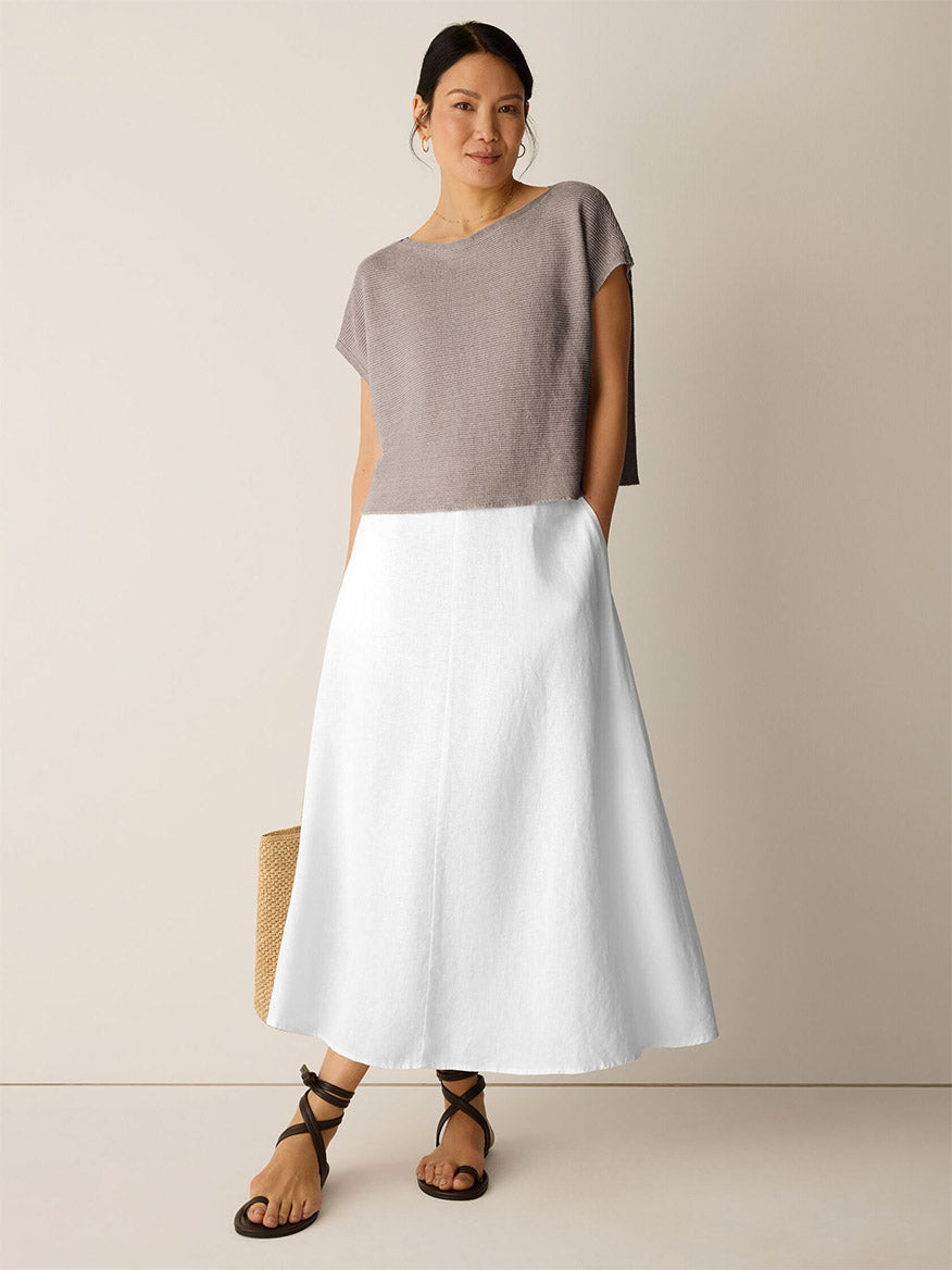 A woman stands against a plain background wearing the Eileen Fisher Organic Linen Délavé Square Top in Walnut, a loose-fitting taupe short-sleeve style, paired with a white A-line midi skirt, black sandals, and holding a woven tote bag.