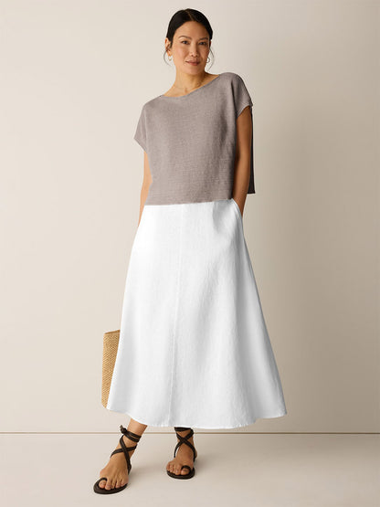 A woman stands against a plain background wearing the Eileen Fisher Organic Linen Délavé Square Top in Walnut, a loose-fitting taupe short-sleeve style, paired with a white A-line midi skirt, black sandals, and holding a woven tote bag.