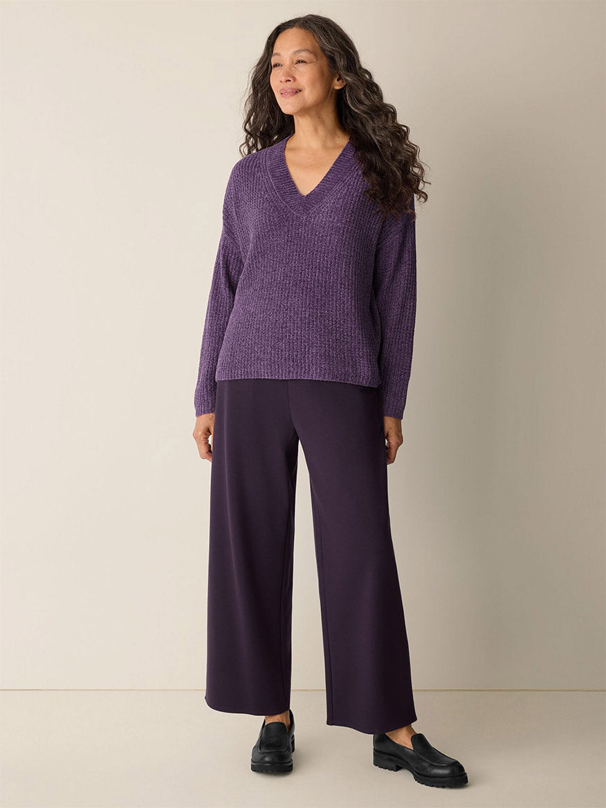 A woman stands against a plain background wearing the Eileen Fisher Chenille Blend V-Neck Top in Mulled Grape, paired with wide-leg dark purple pants and black loafers.