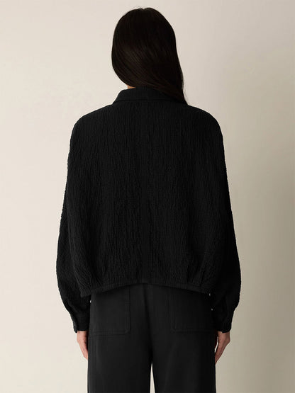 A person with long dark hair is seen from the back against a plain light background, wearing the Eileen Fisher Organic Cotton Doubleweave Classic Collar Jacket in Black paired with black pants.