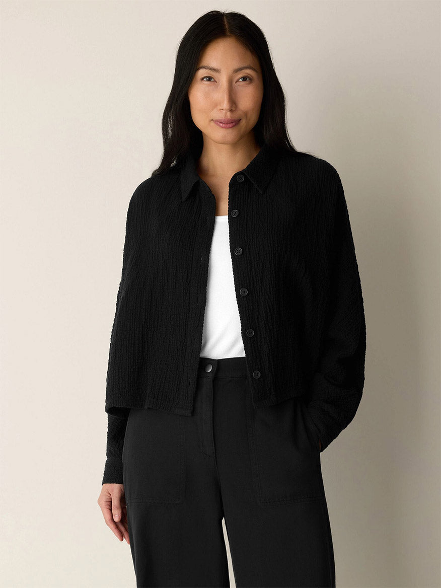 A woman stands against a plain background, wearing the Eileen Fisher Organic Cotton Doubleweave Classic Collar Jacket in Black—a boxy-fit, textured piece—buttoned up over a white top and black pants, with her hands in her pockets.