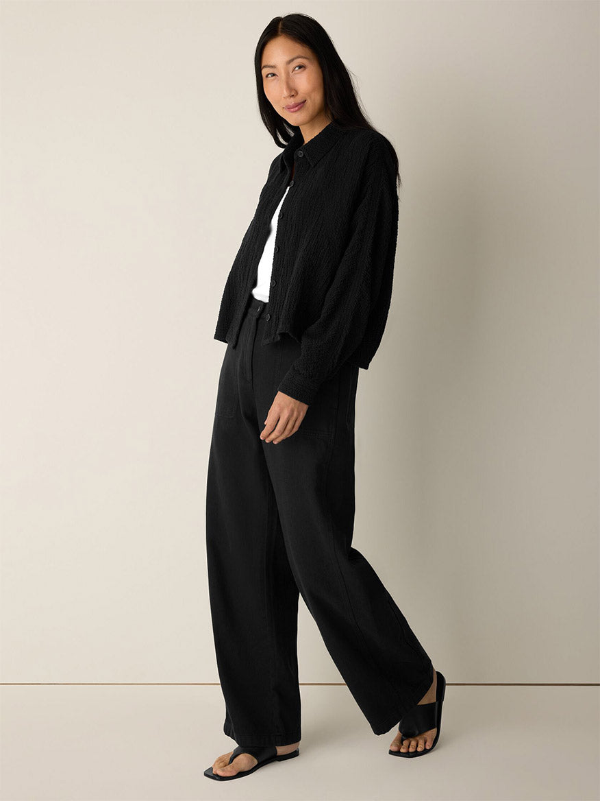 A woman stands against a plain background, wearing the Eileen Fisher Organic Cotton Doubleweave Classic Collar Jacket in Black—a black textured, boxy-fit jacket—paired with a white top, wide-leg black pants, and black sandals.