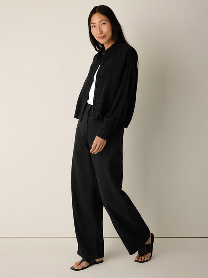 A woman stands against a plain background, wearing the Eileen Fisher Organic Cotton Doubleweave Classic Collar Jacket in Black—a black textured, boxy-fit jacket—paired with a white top, wide-leg black pants, and black sandals.