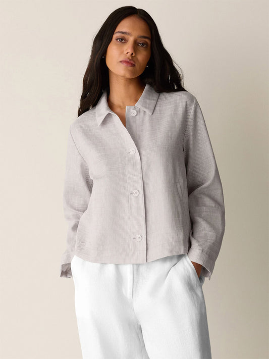 A woman with long dark hair wears the Eileen Fisher Striped Linen Sheen Classic Collar Jacket in Wheat and white pants, standing against a plain light background.