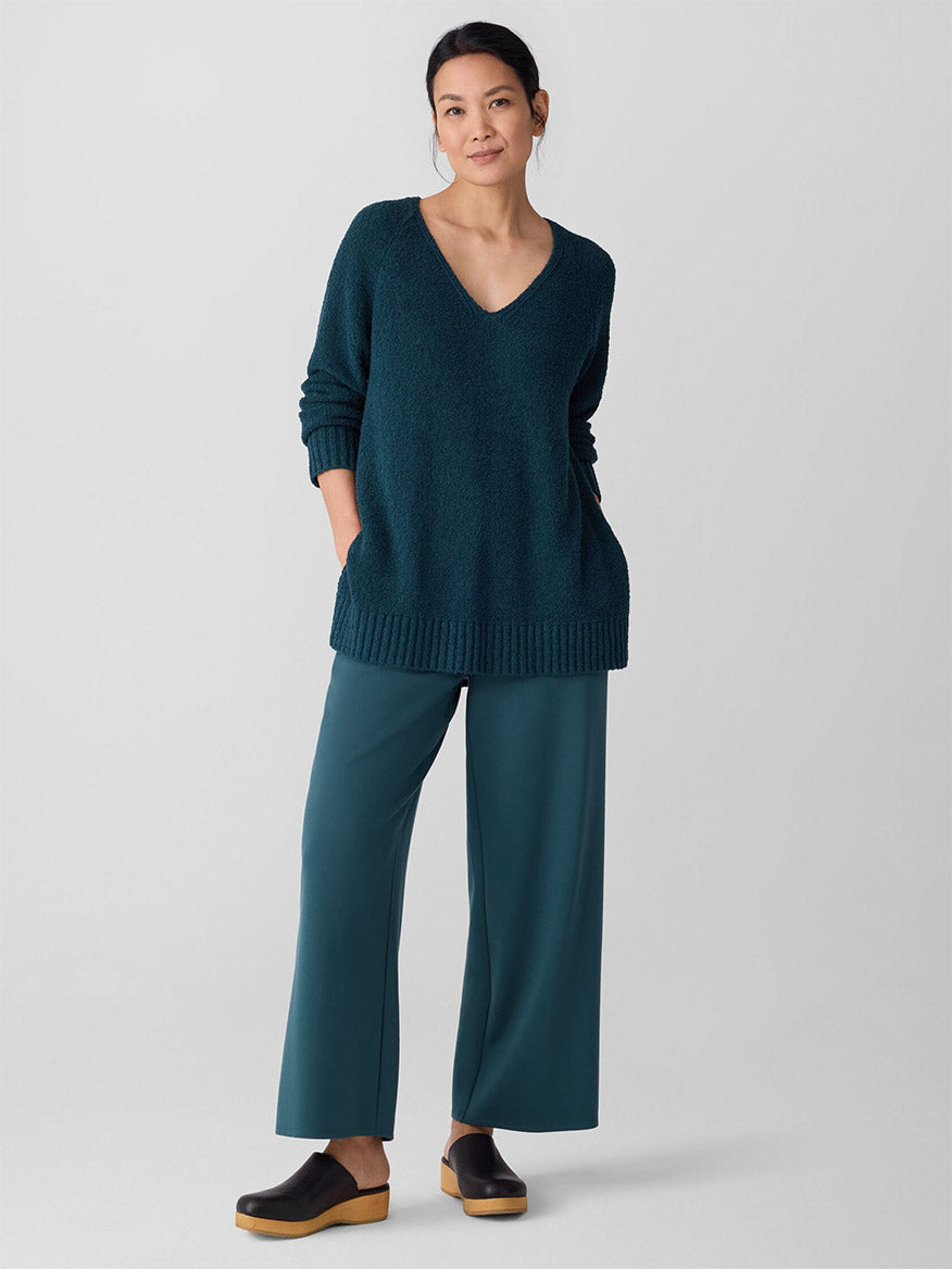 A woman stands against a plain background wearing the Eileen Fisher Cotton Fluff V-Neck Top in Alpine, paired with teal wide-leg pants and black clogs.