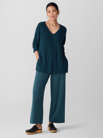 A woman stands against a plain background wearing the Eileen Fisher Cotton Fluff V-Neck Top in Alpine, paired with teal wide-leg pants and black clogs.