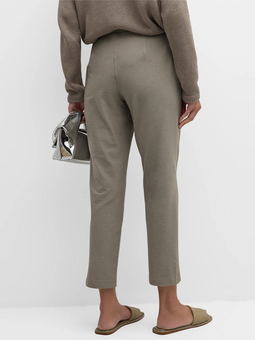 A person is standing, facing away, wearing a beige sweater, Eileen Fisher Washable Stretch Crepe Pants in Dove, and brown slide sandals, holding a metallic silver handbag.