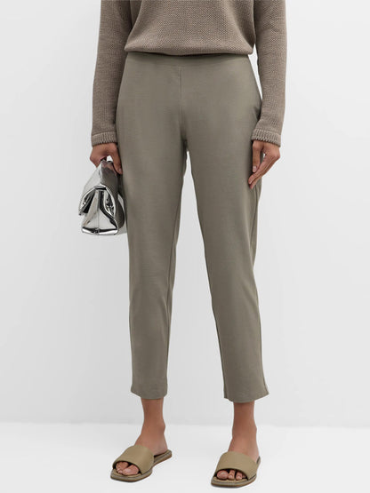 Person wearing a beige sweater, Eileen Fisher Washable Stretch Crepe Pant in Dove, and beige slide sandals, holding a silver handbag.