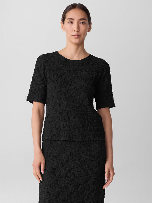 A woman stands facing forward, wearing the Eileen Fisher Bubble Jacquard Crew Neck Top in Black against a plain light background.