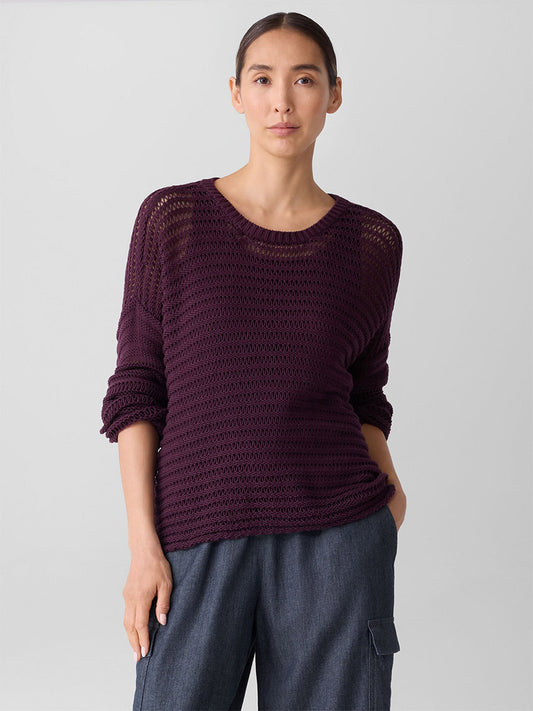 A person wears the Eileen Fisher Peruvian Organic Cotton Cord Crew Neck Top in Blackberry, featuring a loose-knit, boxy fit, paired with blue pants against a plain light gray background.
