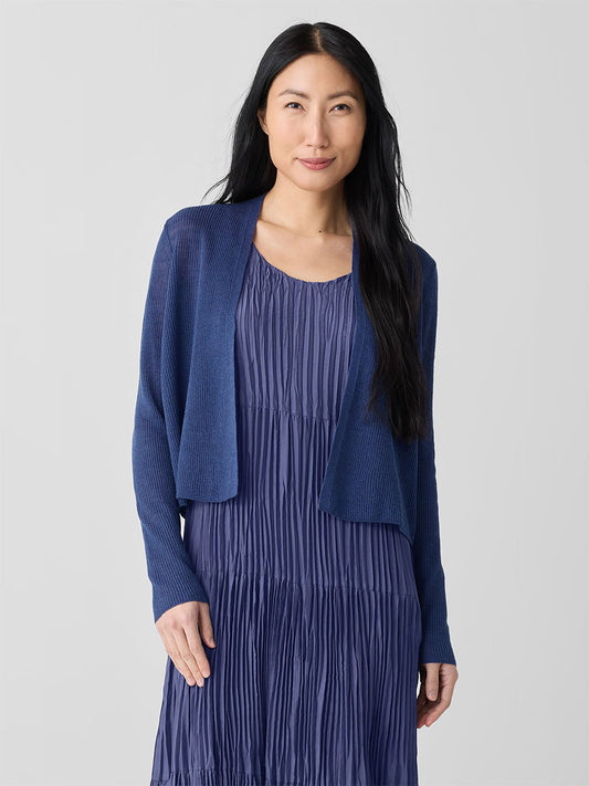 A woman with long black hair stands against a plain background, wearing an Eileen Fisher Organic Linen Cotton Cropped Cardigan in Bluette and a pleated dress.