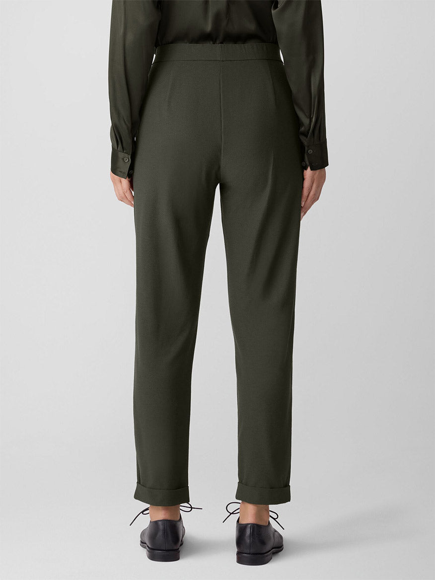 Wearing the Eileen Fisher Washable Stretch Crepe Cuffed Pant in Woodland, paired with a matching long-sleeve top and black shoes, they stand with their back to the camera against a plain light background. The outfit is stylish and easy-care washable.