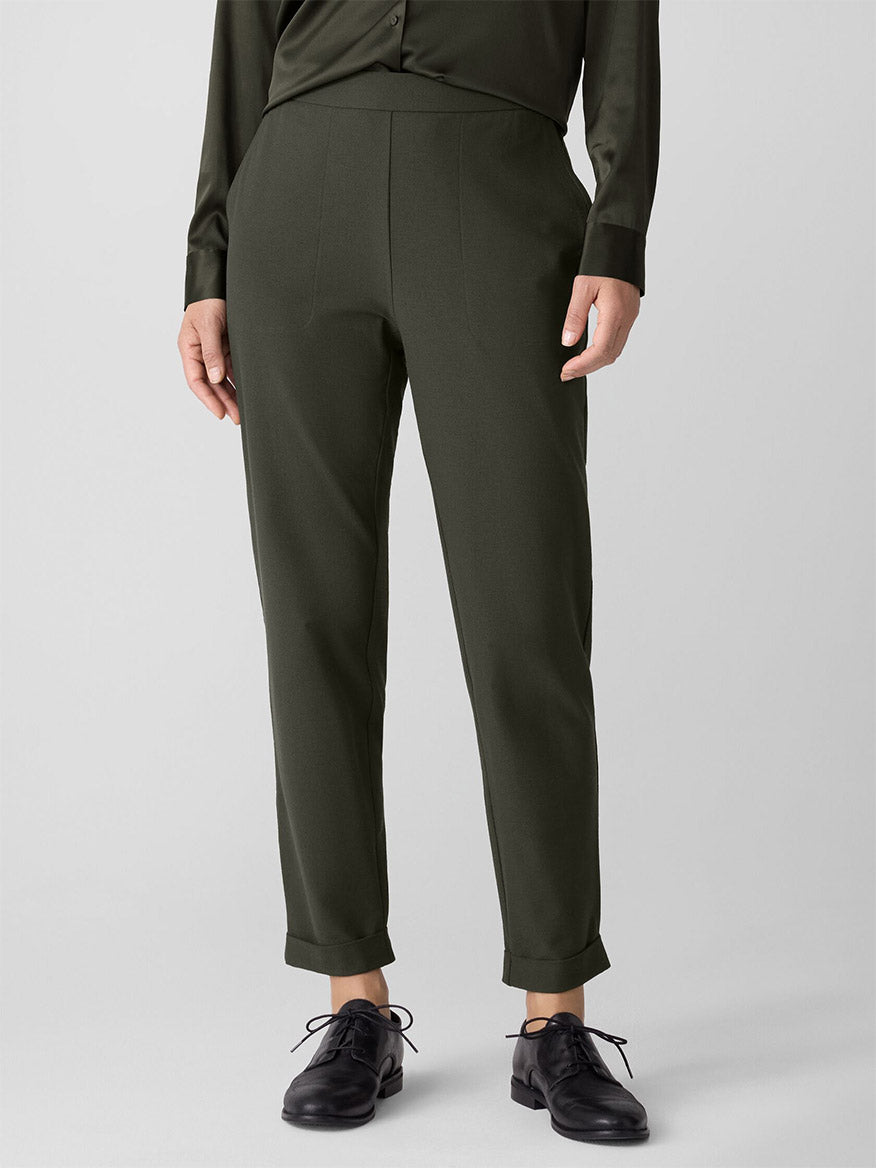 Person wearing Eileen Fisher Washable Stretch Crepe Cuffed Pant in Woodland, a dark green long-sleeve shirt, and black oxford shoes, standing against a plain light gray background.