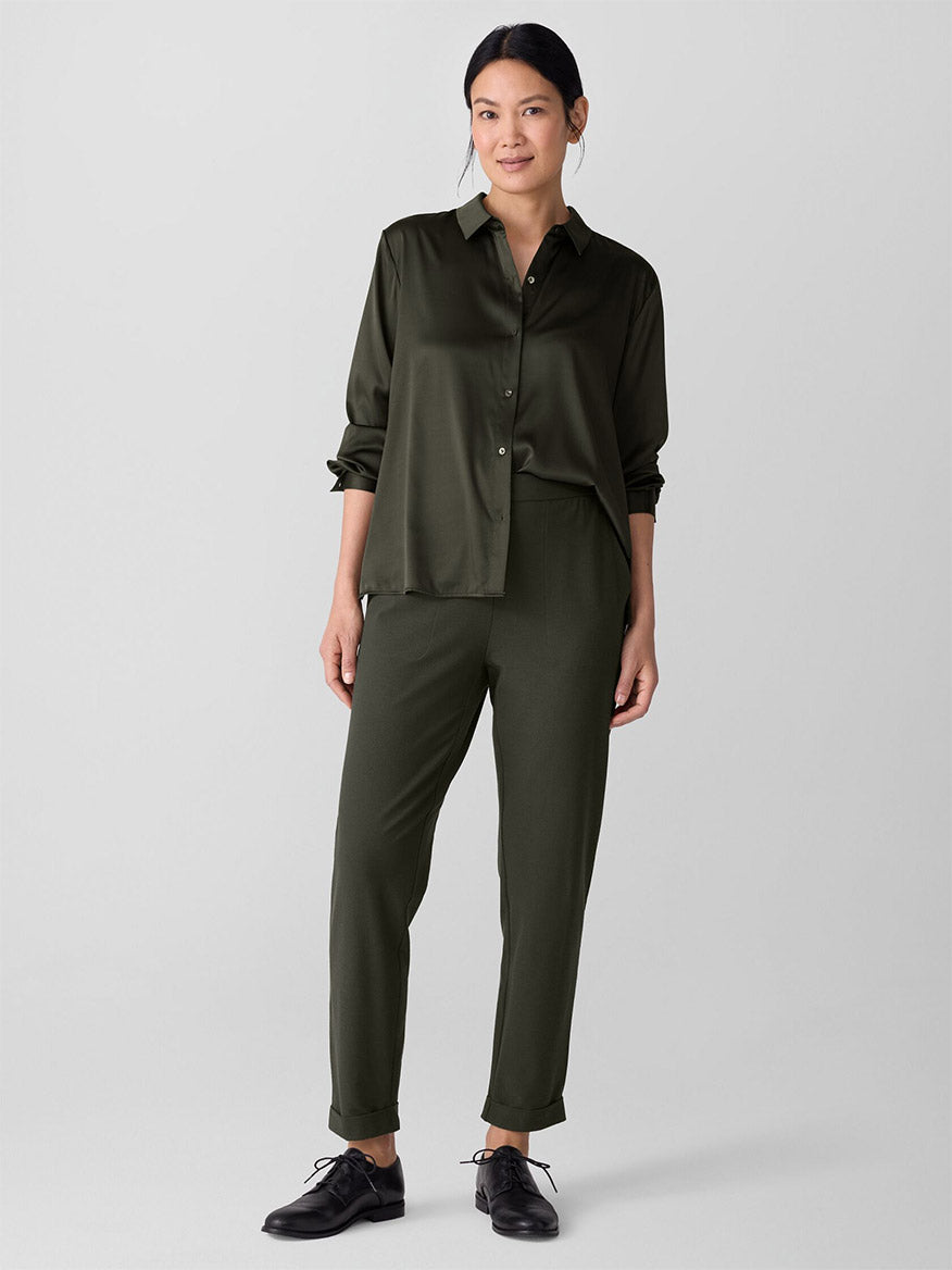 A woman stands against a plain background wearing an olive green button-up shirt, black lace-up shoes, and the Eileen Fisher Washable Stretch Crepe Cuffed Pant in Woodland.
