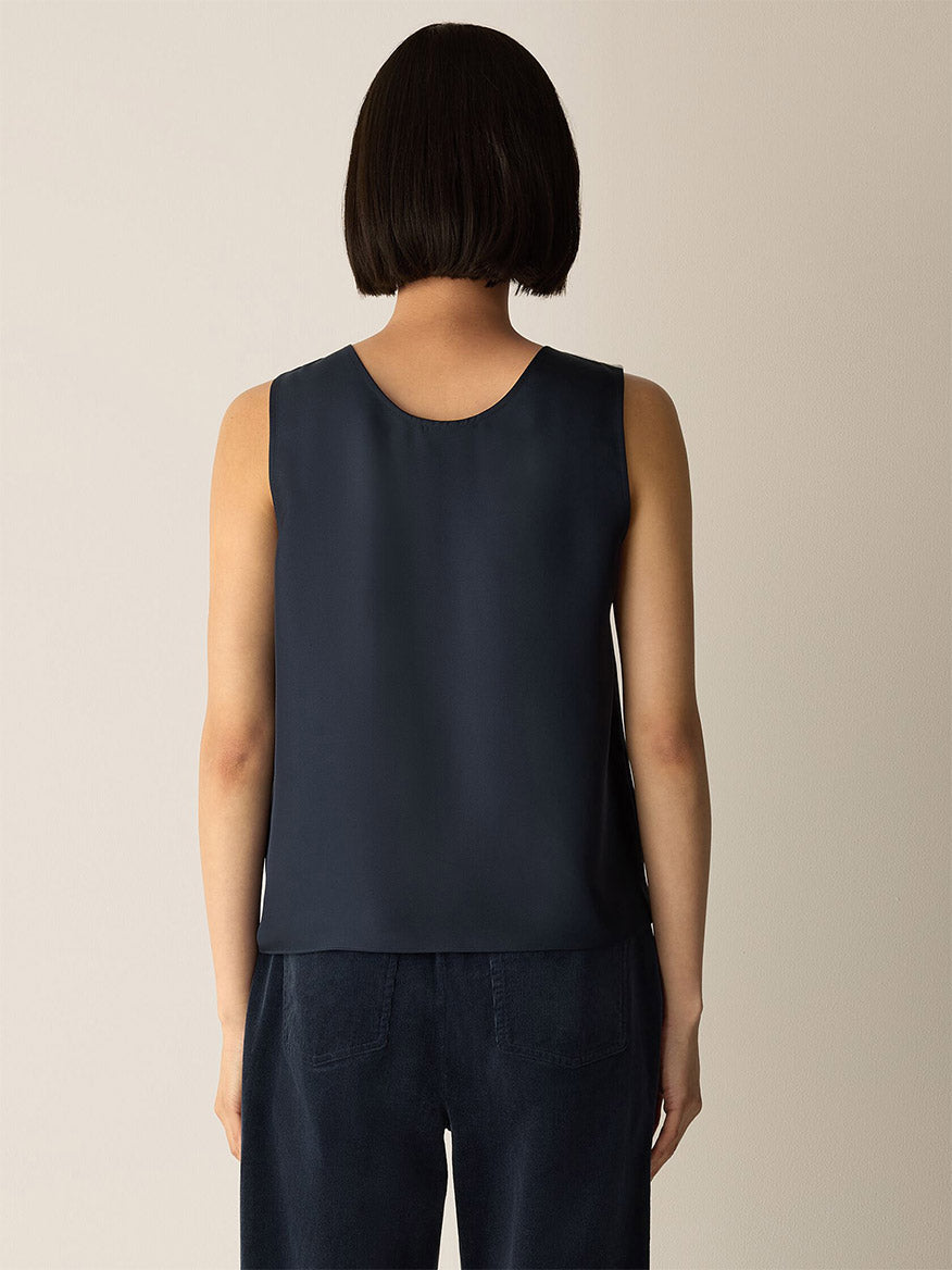 A person with short dark hair stands facing away, wearing the Eileen Fisher Silk Twill Curved V-Neck Tank in Maritime and dark pants against a plain light-colored background.