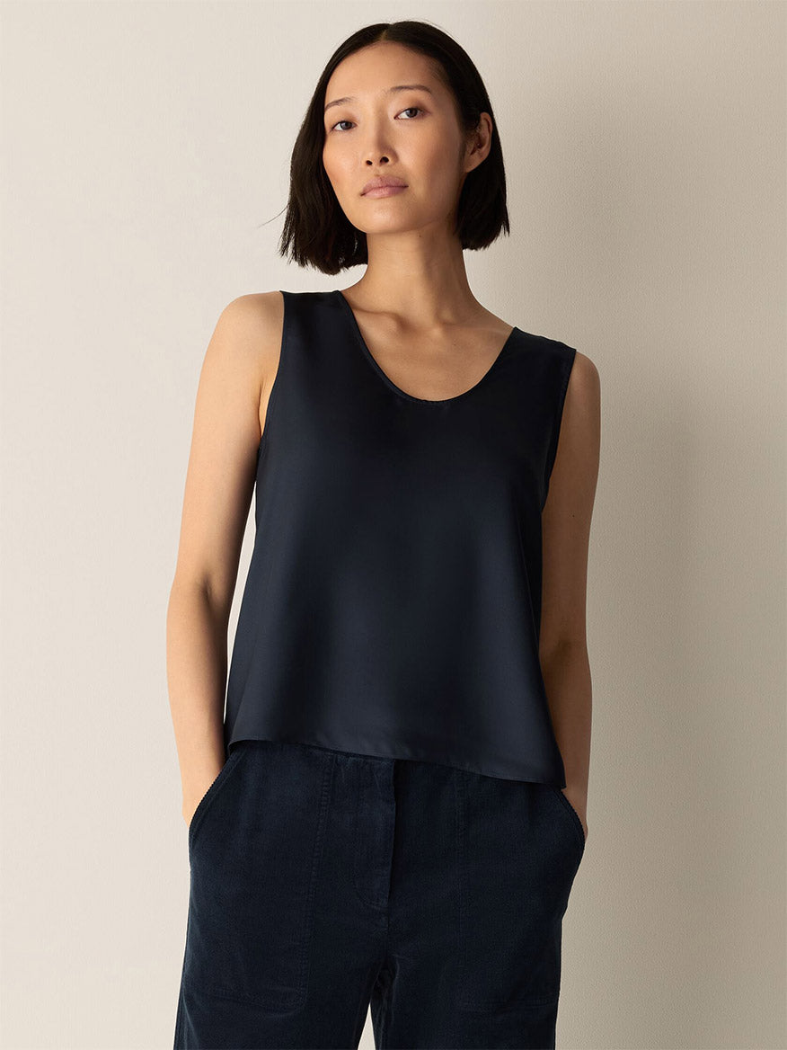 A woman with a short bob hairstyle stands against a plain light-colored background with her hands in her pockets, wearing the Eileen Fisher Silk Twill Curved V-Neck Tank in Maritime and dark pants.