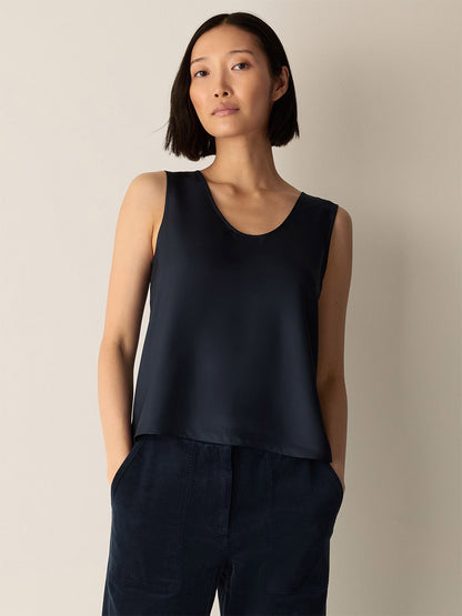 A woman with a short bob hairstyle stands against a plain light-colored background with her hands in her pockets, wearing the Eileen Fisher Silk Twill Curved V-Neck Tank in Maritime and dark pants.