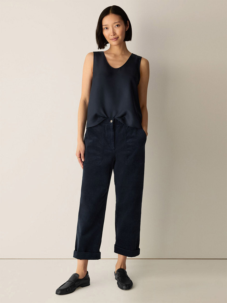 A woman stands against a plain wall wearing the Eileen Fisher Silk Twill Curved V-Neck Tank in Maritime, paired with loose dark trousers with rolled cuffs and black slip-on shoes, her hands in her pockets.