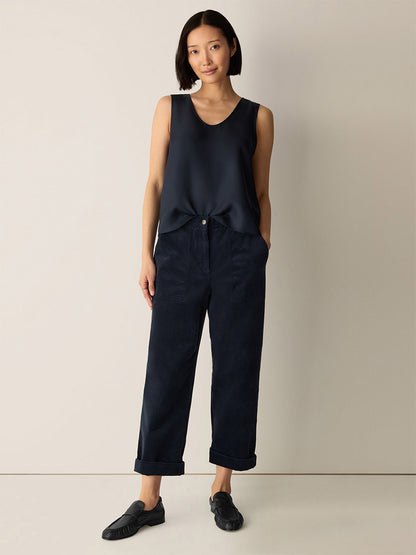 A woman stands against a plain wall wearing the Eileen Fisher Silk Twill Curved V-Neck Tank in Maritime, paired with loose dark trousers with rolled cuffs and black slip-on shoes, her hands in her pockets.
