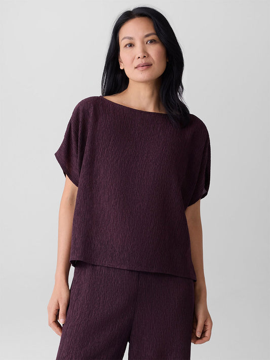 A woman with long dark hair wears the Eileen Fisher Woven Plissé Dolman Sleeve Top in Blackberry, paired with matching pants, standing against a plain light background.