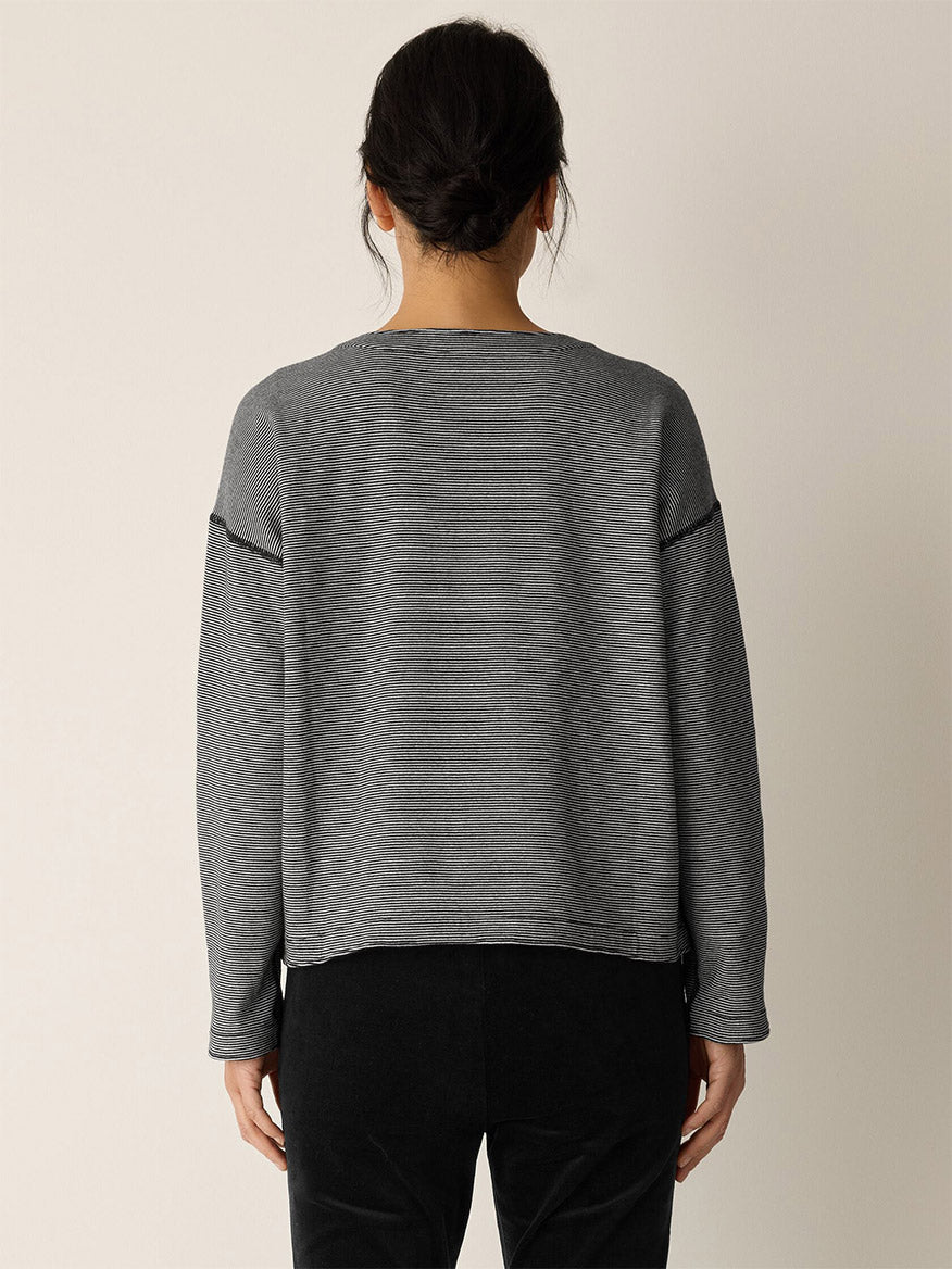 A person with dark hair in a low bun stands with their back to the camera, wearing the Eileen Fisher Doubleknit Organic Cotton Stripe Reversible Top in Black/Ivory and black pants against a plain light background.