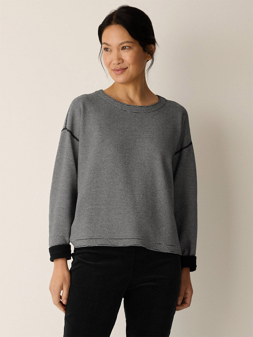 A woman stands against a light background, wearing the Eileen Fisher Doubleknit Organic Cotton Stripe Reversible Top in Black/Ivory and black pants, looking slightly to the side and smiling.