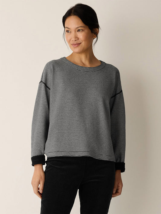 A woman stands against a light background, wearing the Eileen Fisher Doubleknit Organic Cotton Stripe Reversible Top in Black/Ivory and black pants, looking slightly to the side and smiling.
