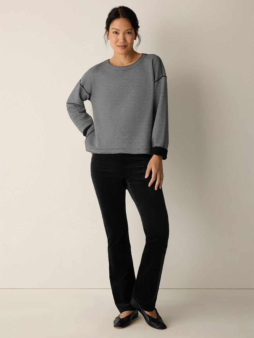 A woman stands against a plain light wall wearing the Eileen Fisher Doubleknit Organic Cotton Stripe Reversible Top in Black/Ivory, paired with black pants and flats, with one hand in her pocket.