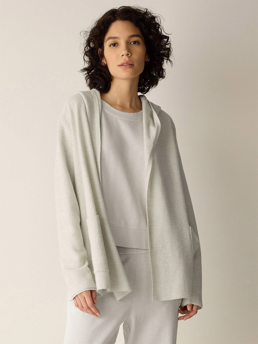 A person wears an open, fine wool hooded cardigan from the Eileen Fisher Fine Felted Wool Hooded Cardigan in Regenerative Wool in Plaster over a matching top.