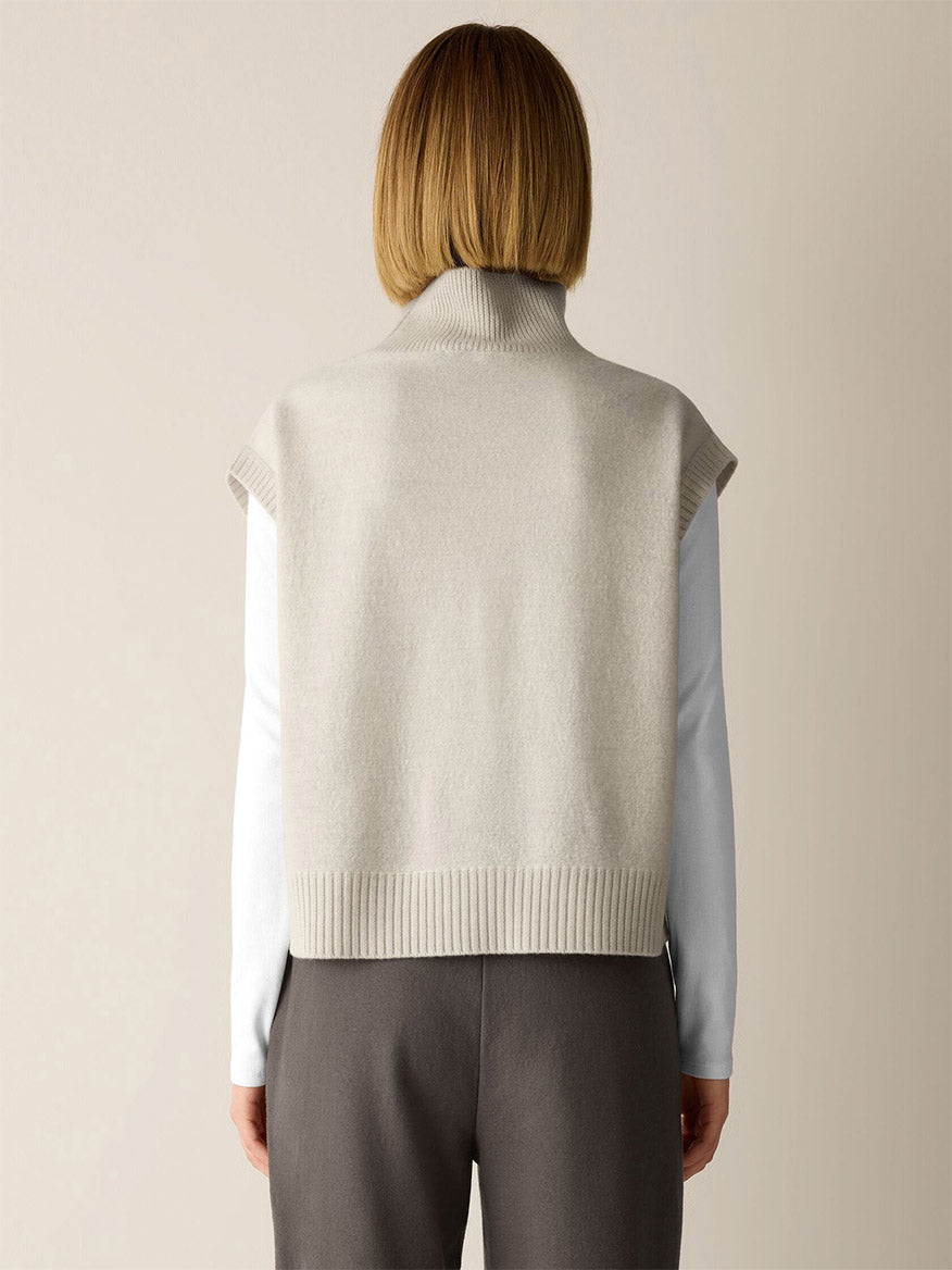 Back view of a person wearing an Eileen Fisher Fine Felted Wool Turtleneck Top in Regenerative Wool in Plaster over a long-sleeved white top and charcoal trousers.