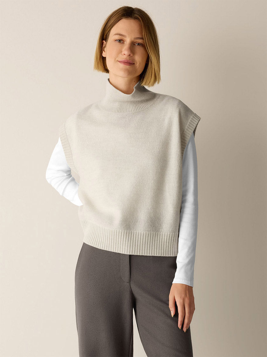A woman wears an Eileen Fisher Fine Felted Wool Turtleneck Top in Regenerative Wool in Plaster over a white long-sleeve shirt and grey trousers.