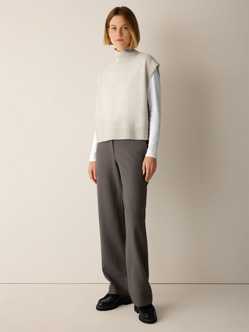 A woman wearing an Eileen Fisher Fine Felted Wool Turtleneck Top in Regenerative Wool in Plaster over a white long-sleeve shirt, paired with gray wide-leg trousers and black loafers.