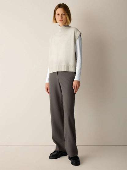 A woman wearing an Eileen Fisher Fine Felted Wool Turtleneck Top in Regenerative Wool in Plaster over a white long-sleeve shirt, paired with gray wide-leg trousers and black loafers.