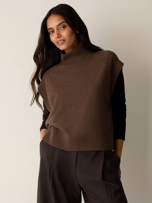 A woman wears an Eileen Fisher Fine Felted Wool Turtleneck Top in Regenerative Wool in Hazelwood, styled as a brown wool turtleneck vest over a black long-sleeved shirt.