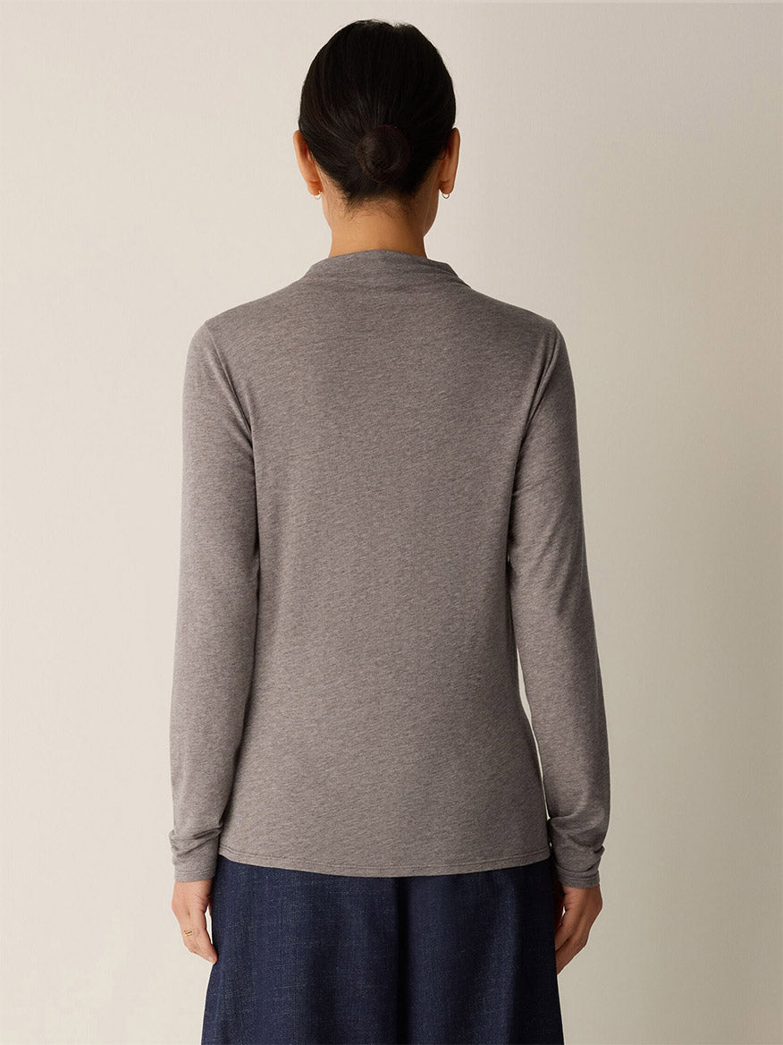 A person stands facing away from the camera, wearing the Eileen Fisher Mélange Sheer Cotton Blend Knit Funnel Neck Top in Cobblestone and a dark blue skirt, with hair tied back.